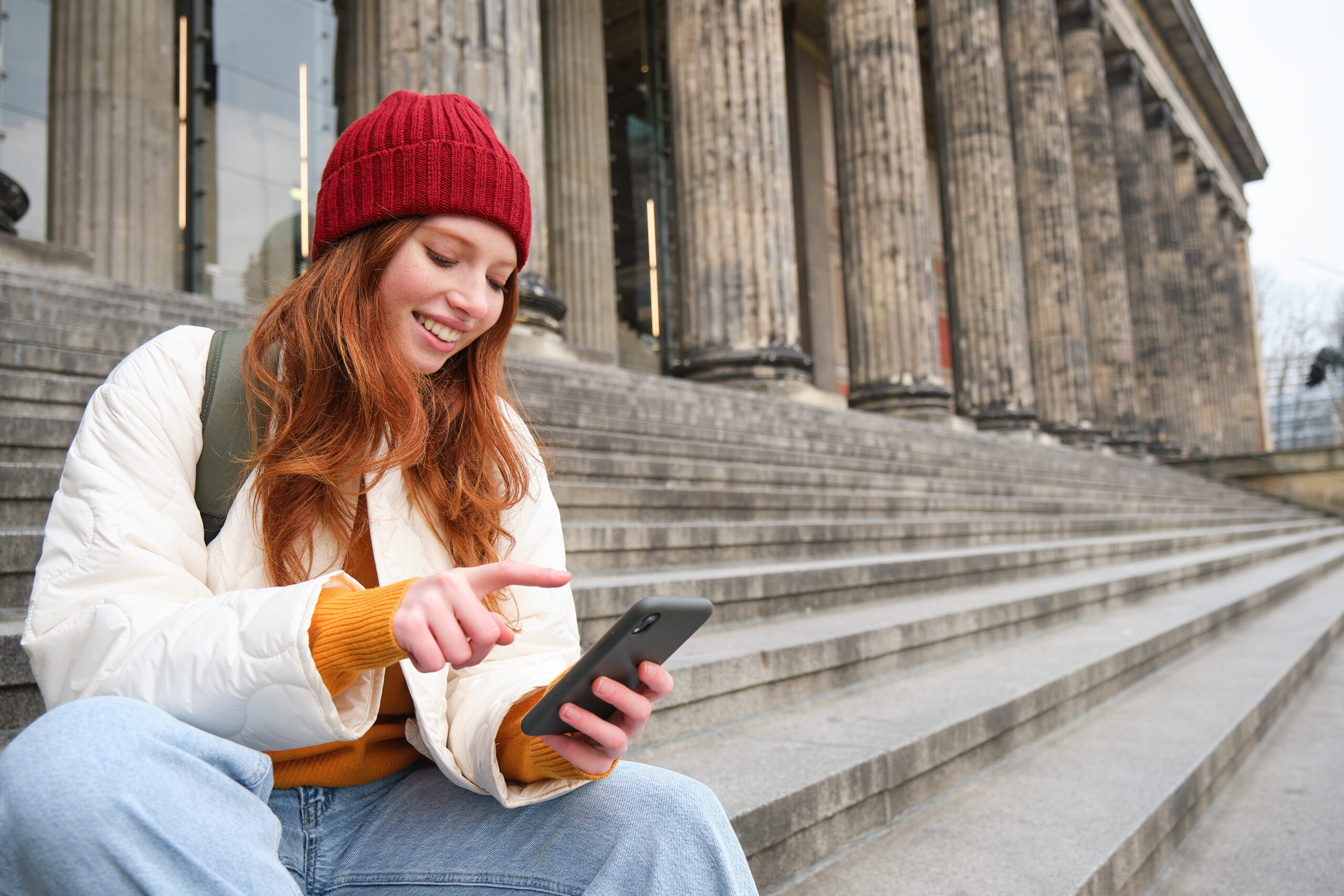 Stylish young redhead woman, talking on mobile phone app, using social media application, looking for something online on smartphone, sits on stairs outdoors.